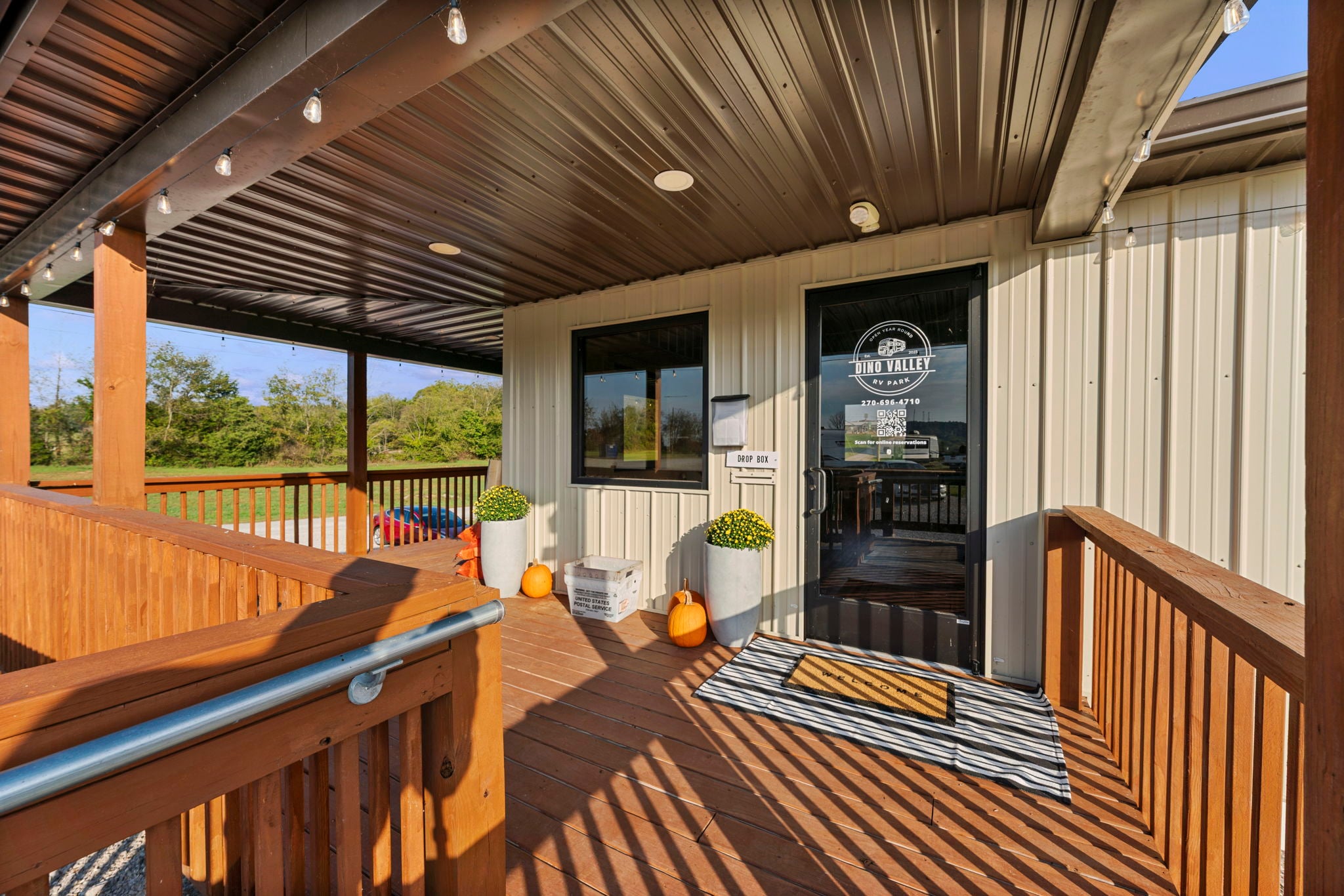 A metal-roofed porch with wooden railings leads to a glass door marked "Tap Truck." Two planters with yellow flowers and three pumpkins decorate the entrance. Trees and blue sky are visible in the background.