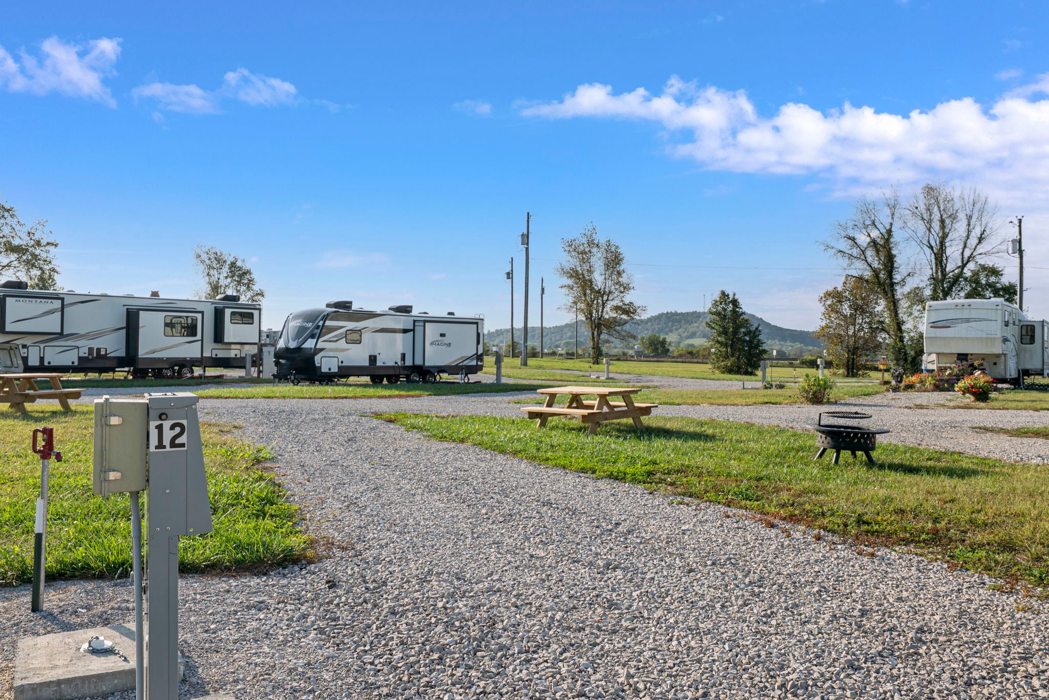 Rows of RVs parked on gravel lots at a campground, with picnic tables, a fire pit, grassy areas, and a blue sky with scattered clouds in the background. A power hookup post labeled "12" is in the foreground.